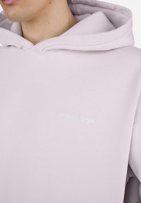 Hoodie lilas clair en tissu doux, doté d'une poche kangourou et d'un petit logo brodé sur la poitrine.