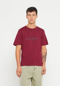Burgundy színű pamut póló rövid ujjakkal, kerek nyakkivágással, elöl fekete írásos logónyomattal. A modellen világos színű nadrág van.