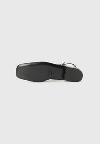 Chaussure à talon haut noir avec une surface lisse et brillante et un bout arrondi. La semelle est plate avec une légère texture de traction et une marque en relief.