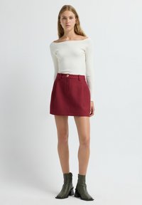 Witte geribbelde off-shoulder top gecombineerd met een rode gestructureerde minirok met een voorknoop en zakken. Model draagt groene enkellaarsjes.