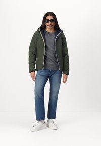 Grøn quiltet jakke med en marineblå hætte, grå skjorte under, blå jeans og hvide sneakers. Har en lynlås og stribede detaljer.