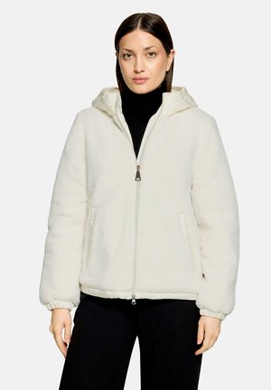Frau trägt eine weiße Fleecejacke mit Reißverschluss und Kapuze über einem schwarzen Rollkragenpullover und schwarzen Hosen, stehend vor einem einfarbigen Hintergrund.