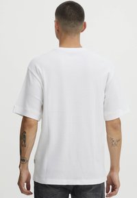 Hvid kortærmet t-shirt med en tekstureret, ribbet stof. Den har en rund halsudskæring og op rullede ærmer, båret af en person med tatoveringer på armen.