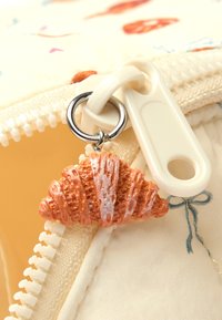 Zipper pull z teksturowanym, pomarańczowym zawieszką w kształcie rogalika, przymocowanym do białego zamka błyskawicznego, na tle wzorzystej, kremowej tkaniny.