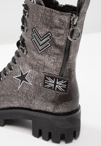 Botte de combat grise avec patchs en étoile noire, chevron et Union Jack, fermeture éclair noire avec tirette ronde, et semelle épaisse à crampons.
