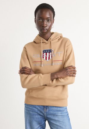 Beige hettegenser med snor, med en frontlogo i blått, rødt og hvitt. Modellen kombinerer den med blå jeans. Myk stoffkvalitet.