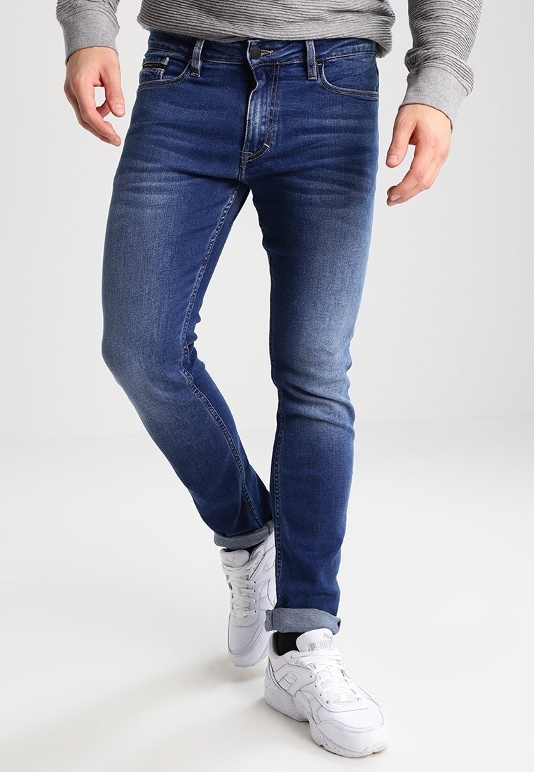 Jeans de mezclilla azul oscuro con corte slim, que presentan un lavado desgastado, diseño de cinco bolsillos y puños enrollados, combinados con zapatillas deportivas blancas.