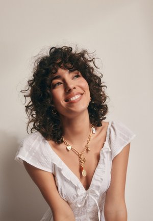 Zlatý náhrdelník s různými přívěsky visí na ženě, která má na sobě bílou, texturovanou blůzu s volánkovými rukávy a výstřihem do V.