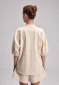 Beige Bluse mit gestickten Details, Ballonärmeln, floralem Muster, rundem Ausschnitt und lockerer Passform. Glatte Textur mit Seitenschlitzdetails.