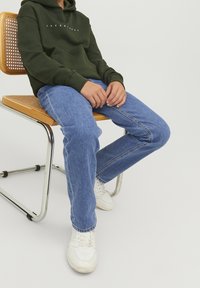 Grön hoodie med framficka och logotyp, blå denimjeans och vita sneakers, sittandes på en stol av trä och metall mot en vit bakgrund.