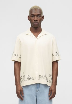 Homme aux cheveux blonds courts portant une chemise crème à boutons avec des croquis noirs de désert et de palmiers, associée à un jean bleu clair.