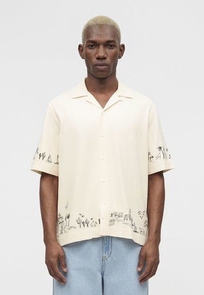 Hombre con el pelo corto y rubio que lleva una camisa color crema con botones y dibujos negros de desiertos y palmeras, combinada con vaqueros azul claro.