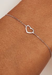 Zilveren kettingarmband met een minimalistische hartbedel, op een gebruinde huid achtergrond, die het delicate ontwerp benadrukt.