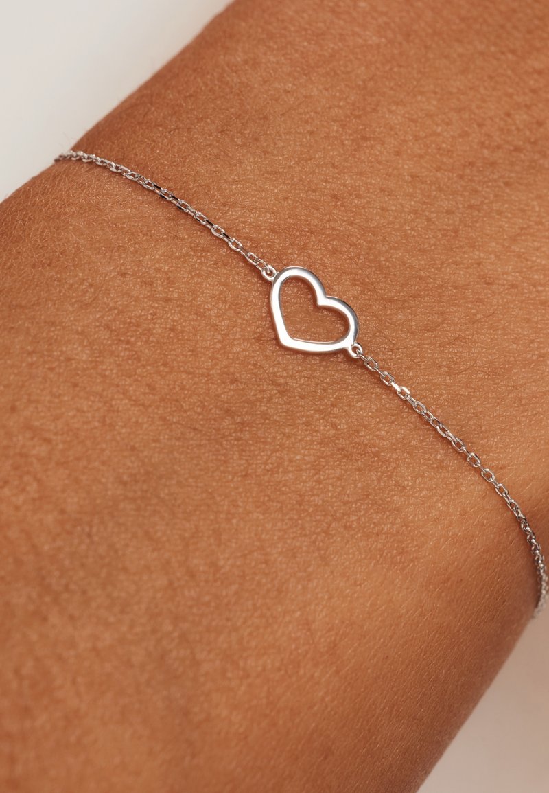 Zilveren kettingarmband met een minimalistische hartbedel, op een gebruinde huid achtergrond, die het delicate ontwerp benadrukt.