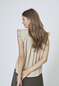 Beige ärmelloses Top mit rüschengesäumten Schultern, aus leichtem Stoff gefertigt. Bietet eine lockere Passform und einen abgerundeten Saum für zusätzlichen Komfort.