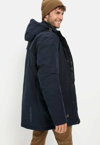 Marineblaue isolierte Parka mit Kapuze, verfügt über einen Reißverschluss und Druckknopfverschluss, Seitentaschen und ein Logo auf der linken Seite. Braune Mütze.