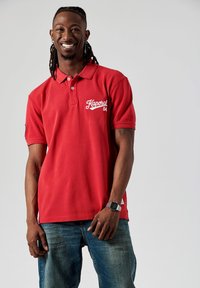 Rode polo met korte mouwen, voorzien van een wit logo en nummer op de borst. Gecombineerd met blauwe jeans. Eenvoudig, casual ontwerp.