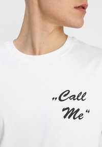 Witte katoenen t-shirt met zwarte opdruk met de tekst "Bel me" in een cursieve lettertype, met een ronde halslijn en korte mouwen.