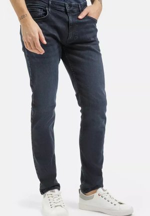 Mężczyzna w ciemnych dopasowanych dżinsach, z jedną ręką w kieszeni i białych trampkach, na jednolitym jasnym tle.