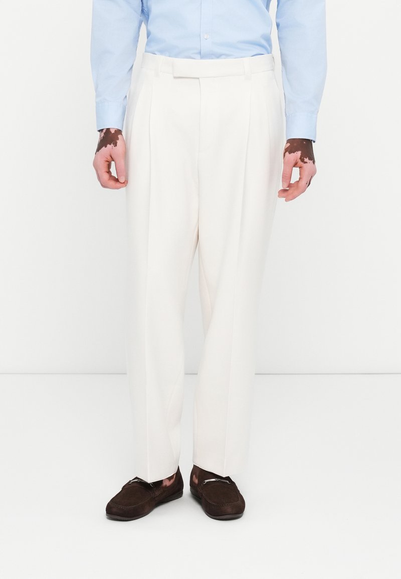 Witte op maat gemaakte broek met een geplooide voorkant, gemaakt van een gladdige stof, gecombineerd met donkerbruine instapchoenen en een lichtblauw overhemd met kraag.