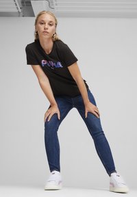 Fekete póló színes grafikával az elején, kék szűk farmerrel és fehér sneakers-sel, rózsaszín akcentussal.