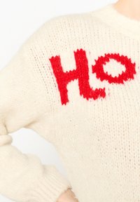 Kreminė megztinė su raudonu „Ho Ho“ išsiuvinėtu užrašu, pasižyminti laisvu kirpimu, raukšlėtais rankogaliais ir tekstūruotu audiniu.