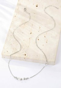 Collana d'argento con una delicata catena, caratterizzata da tre perle e motivi a stella. Adagiata su una superficie di pietra chiara e texture.