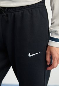 Fekete melegítőnadrág, puha anyagból készült, rugalmas derékkal, oldalzsebekkel és a bal combon hímzett fehér Nike logóval.