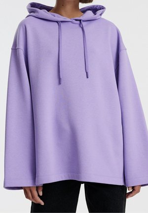 Person wearing oversized lavender hoodie with drawstrings and long sleeves, paired with black pants, against plain light background.