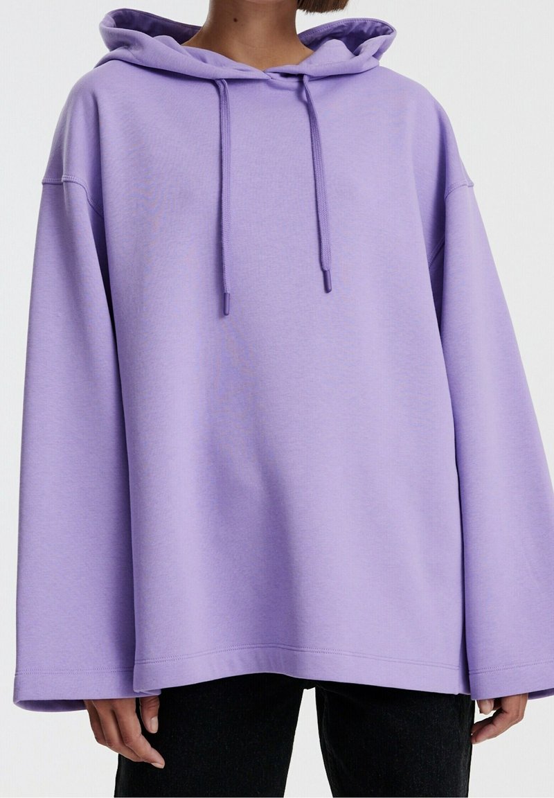 Personne portant un sweat à capuche lavande oversized avec cordons et manches longues, associé à un pantalon noir, sur un fond clair uni.