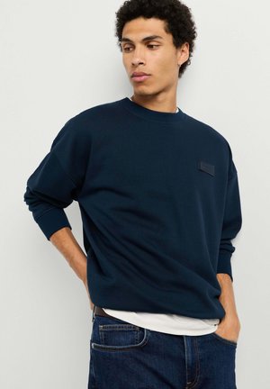 Junger Mann trägt einen dunkelblauen Pullover und dunkelblaue Jeans, steht mit einer Hand in der Tasche und schaut nach rechts.