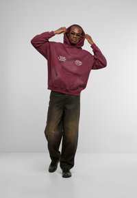 Felpa oversize color marsala con tasca frontale, testo grafico sul petto, abbinata a pantaloni larghi neri e scarpe nere, indossata da un modello.