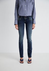 Tamsiai mėlynos džinsinės kelnės su slim fit kirpimu, turinčios išblukusių detalių ir daugiapakopę priekinę užsegimą, derinamos su dryžuota marškinių ir purpurinėmis šlepetėmis.
