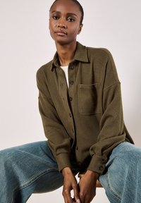 Olive green ribbed cardigan with a shirt collar, front pocket, and brown buttons, paired with loose-fitting light blue jeans.