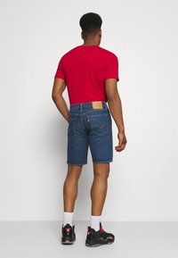 Modré džínové šortky se rovné střihy, koženou nášivkou na zadním pase, nošené s červeným tričkem a černými sportovními botami s akcenty.