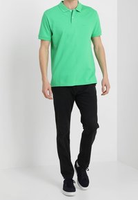 Grön bomullspikétröja med krage, två knappar och en broderad logotyp. Matchas med svarta jeans och marinblå sneakers.