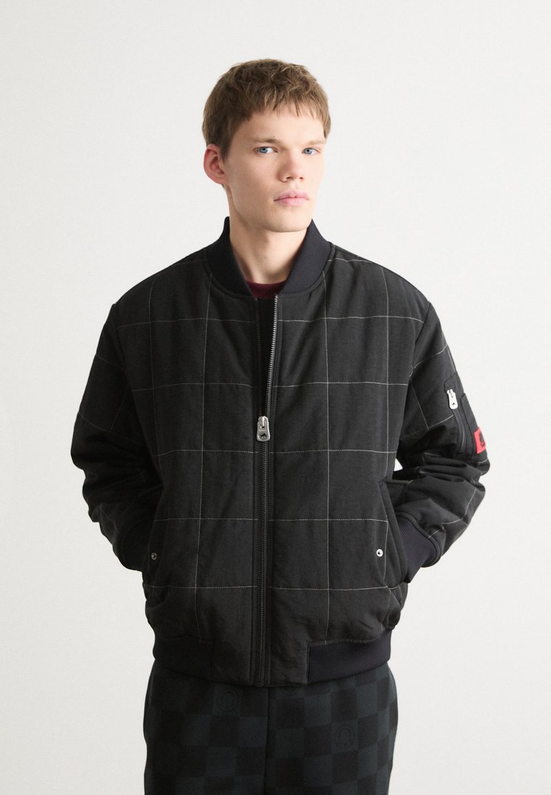 Veste bomber noire avec motif à carreaux, tissu épais, fermeture éclair, poignets côtelés et un logo rouge en accent sur la manche.