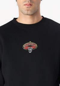 Black sweatshirt with a round neckline featuring an embroidered design of a skull wearing a sombrero, accented with red and yellow details.