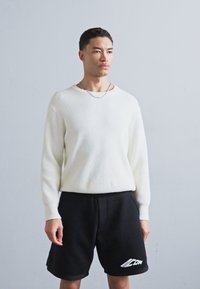 Suéter blanco de punto acanalado y pantalones cortos negros con un logotipo gráfico. El suéter cuenta con mangas largas; los pantalones cortos tienen un corte relajado y bolsillos laterales.