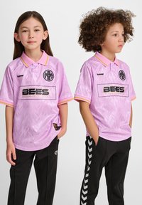 Rosa sporttröjor med ett geometriskt mönster, krage och varumärke. I kombination med svarta byxor prydda med ett vitt zigzag-mönster.