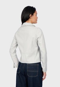 Weißes Lederjackett mit tailliertem Schnitt, langen Ärmeln und eingekerbtem Kragen. Über dunklen Denim-Hosen getragen, die eine glatte Textur zeigen.