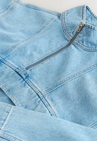 Veste en denim bleu clair avec une fermeture éclair argentée à l'avant et un col montant, mettant en valeur les coutures détaillées et la texture du tissu.