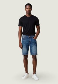 Joven de pie con camiseta negra, pantalones cortos vaqueros azules y zapatillas blancas, con una mano en el bolsillo y fondo liso.