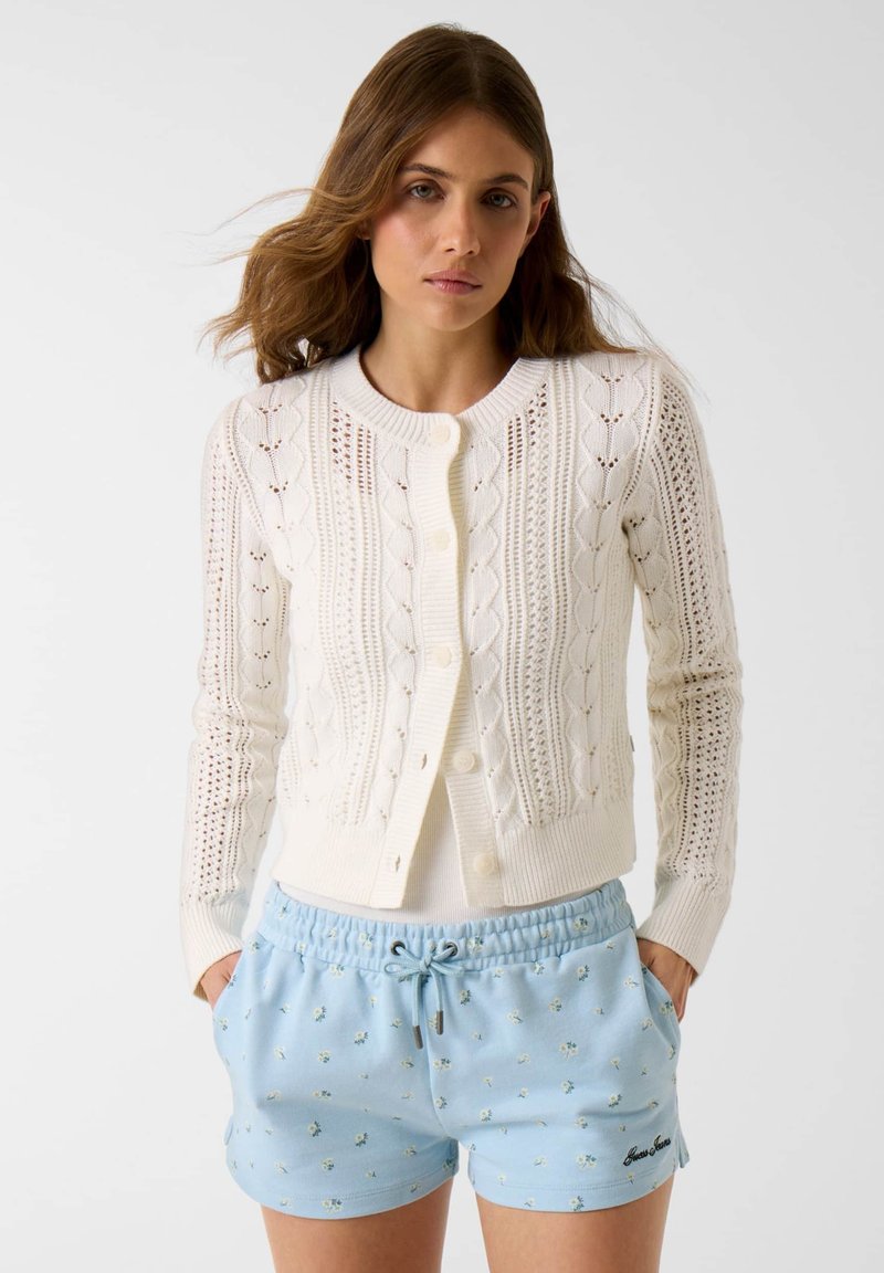Mladá žena v bielej pletenej zapínacej kardigáne a svetlomodrých kvetovaných šortkách so sťahovacou šnúrkou, stojaca s rukami v vreckách.