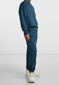 Person som bär en blågrön sweatshirt och matchande joggers med vita klumpiga sneakers, stående mot en enfärgad ljus bakgrund.