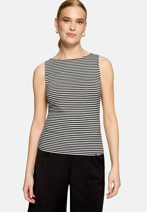 Femme aux cheveux blonds portant des boucles d'oreilles créoles dorées et un haut sans manches à rayures horizontales noires et blanches avec un pantalon noir.