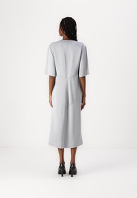 Graues Midikleid mit kurzen Ärmeln, geradem Schnitt, strukturiertem Stoff und einer subtilen Naht an der Rückseite. Schwarze hochhackige Schuhe getragen.