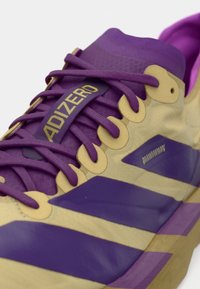 Chaussure de sport violette et crème avec une tige texturée, des lacets violets et un logo doré. Présente des rayures violettes bien visibles et des accents de matériel élégants.