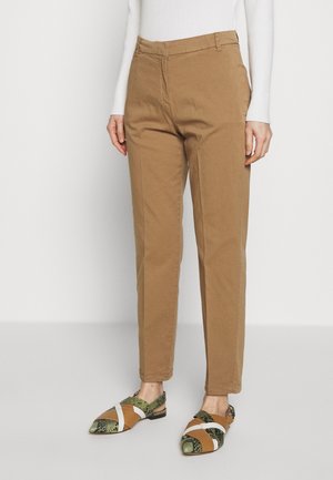 Kvinde iført tanfarvede bukser med lige ben og høj talje samt mønstrede flade sko, stående mod en hvid baggrund.