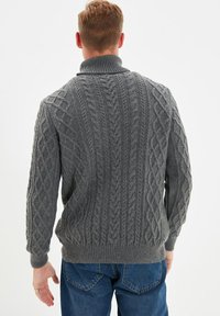 Grau gestrickter Pullover mit Zopfmuster, hohem Kragen und gerippten Bündchen, in lockerer Passform. Kombiniert mit blauen Jeans.